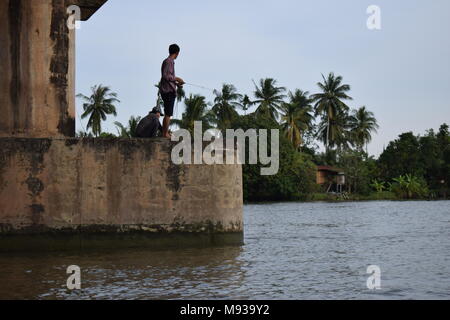 2 fishermans Angeln unter einer großen Brücke über den Fluss Mae Klong in Thailand von einem Touristenboot tour mit blauem Himmel und hohen Palmen. Stockfoto