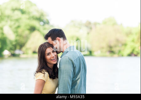 Fröhliche und spontane Paar in Liebe, halten sich an den Händen auf einem steinernen Pier auf einem natürlichen See Spaß, küssen, umarmen und Lachen im Freien. Stockfoto