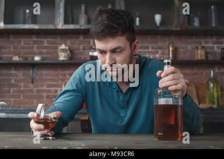 Junger Mann Alkoholsucht Probleme am Tisch sitzen halten Whiskey Flasche und Glas nach unten schaut gleichgültig Stockfoto