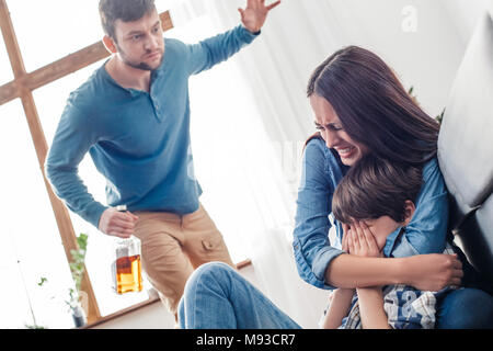 Familie soziale Probleme häuslicher Gewalt Konzept Mann wütend Holding Flasche Whiskey schlagen Frau weinend umarmen Sohn erschrocken close-up Stockfoto