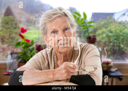 Lächelnd Senior, grauhaarige Frau, holdin Gläser, an der Kamera suchen, in Ihrem Wohnzimmer, vor der Fenster in den Garten. Stockfoto
