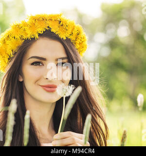 Lächelnde Frau mit einem Löwenzahn Blume auf grünem Gras. Happy Brunette Modell tragen gelbe Blumen Kranz im Freien Stockfoto