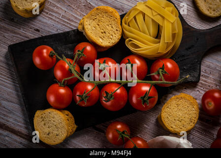 Horizontale Foto mit Top Aussicht auf Zweig mit roten Kirschtomaten, die auf altem Schneidebrett und Holztisch mit Vintage Farbe platziert werden. Bruschetta Stockfoto