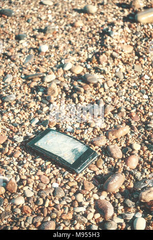 Gebrochene Handy mit gebrochenen Bildschirm auf einem steinigen Strand, selektiver Fokus, Farbe getonte Bild. Stockfoto