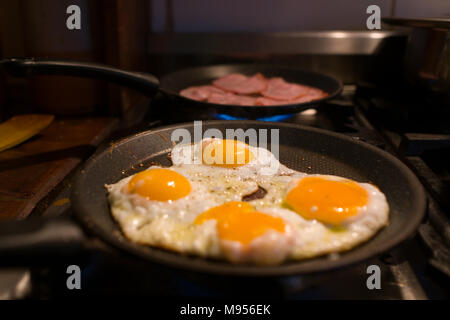 Vier frische Bio Eier braten in einer kleinen Pfanne mit Speck Braten in einer anderen Pfanne im Hintergrund Stockfoto