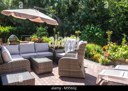 Moderne Rattan Gartenmöbel mit Esstisch, Stühlen und Sonnenschirm im Hintergrund alle von viel Grün und Blumen umgeben Stockfoto