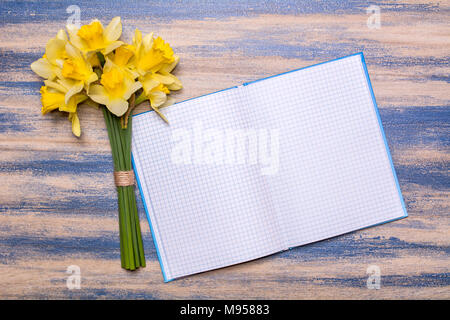 Notizblock auf einem Holzbrett. Gelbe Narzissen Blumen auf einem Holztisch. Blühten blauer Farbe. Hintergrund mit Platz für Text. Ansicht von oben, flach liegen d Stockfoto