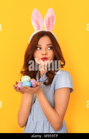 Junge Frau Studio auf gelbe Wand tragen cute dress und hasenohren Holding bunte Eier in Palms schließen isoliert - bis zur Seite schauen neugierig Stockfoto