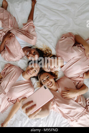 Blick von oben auf die Braut unter selfie mit brautjungfern beim liegen auf dem Bett. Gruppe von Frauen mit Schlafrock liegend auf Bett und Selbstportrait. Stockfoto