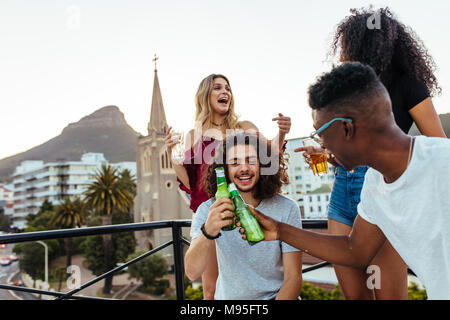 Glückliche junge Männer toasten Biere mit Mädchen lachen und genießen auf der Rückseite auf der Terrasse. Eine Gruppe von Freunden, die auf der Dachterrasse Partei mit Getränken. Stockfoto