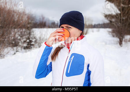 Junge Frau im Winter Sport Jacke trinken heißen Tee von orange Thermoskanne Schale auf kalten bewölkten Tag mit Schnee bedeckt die Landschaft im Hintergrund. Stockfoto