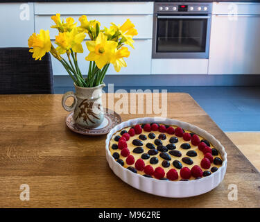 Küche Szene - Narzissen in einer Töpferei Vase und leckeren Käsekuchen In weißen Teller mit Trauben & Berry Topping auf hölzernen Tisch Stockfoto