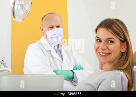 Attraktive Patientin lächelnd in die Kamera im Zahnarztstuhl, reife männlicher Arzt mit Maske im Hinter Stockfoto