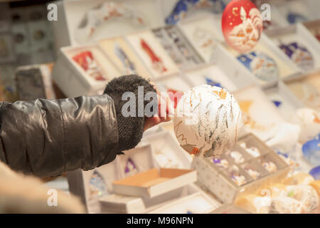 Anbieter hält dekorativen Weihnachtsbaumball in der Hand und macht das Angebot an die Kunden, Weihnachtsmarkt am Abend mit Ambient Kioskbeleuchtung. Stockfoto