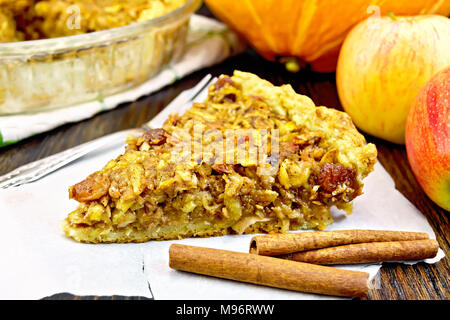 Stück Kuchen mit Äpfeln, Kürbis, Rosinen und Nüssen auf Pergament, Zimt auf dem Hintergrund einer Holzplatte Stockfoto