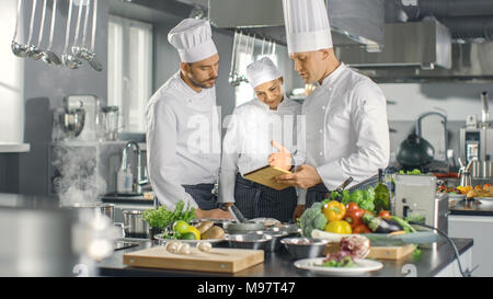 In der modernen Küche Team von Köchen mit Tablet-PC für Rezepte, Sie Lächeln und Diskussion. Küche ist voll von Lebensmittelzutaten, Gemüse. Stockfoto