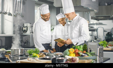In der modernen Küche Team von Köchen mit Tablet-PC für Rezepte, Sie Lächeln und Diskussion. Küche ist voll von Lebensmittelzutaten, Gemüse. Stockfoto