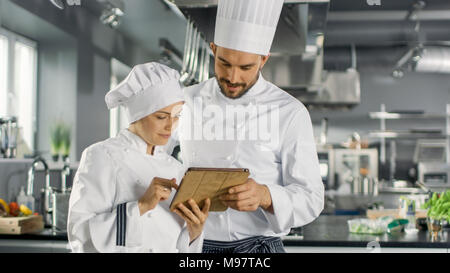 Männliche und weibliche Berühmte Köche sprechen über Ihre Video Blog während der Verwendung von Tablet Computer. Sie arbeiten auf einem großen Restaurant Edelstahl professionelle Küche. Stockfoto