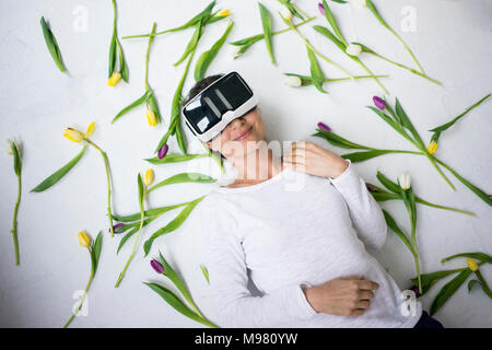 Lächelnde Frau mit VR-Brille lag auf dem Boden inmitten Tulpen Stockfoto