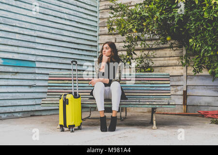 Junge Frau mit gelben Trolley Tasche wartet auf einer Bank Stockfoto