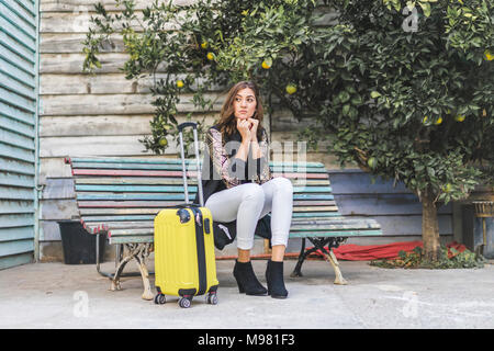Junge Frau mit einem gelben Trolley Tasche wartet auf einer Bank Stockfoto