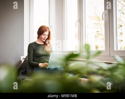 Rothaarige Frau sitzt neben Fenster mit Laptop Stockfoto