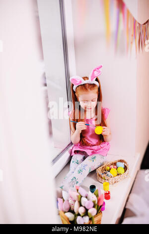 Ein kleines hübsches Mädchen mit roten Haaren mit Hase Ohren auf dem Kopf ist Malen Eier für ein Ostern Korb auf der Fensterbank gegen das Fenster und Ostern d Stockfoto