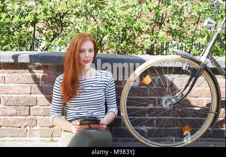 Porträt der rothaarige Frau mit Tablet und Fahrrad sitzen auf einer Mauer Stockfoto