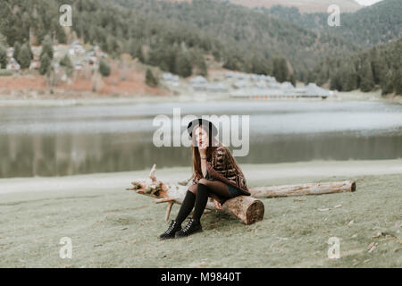 Modische junge Frau mit Hut und Poncho sitzen auf Log auf einer Wiese Stockfoto