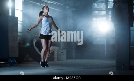 Athletische schöne Frau Übungen mit Jump/Springseil in ein Fitnessstudio. Sie ist in Schweiß von der intensiven Cross Fitness Training abgedeckt. Dunkle Atmosphäre Stockfoto