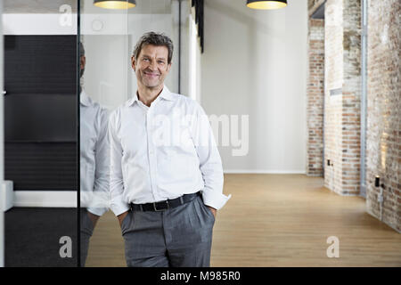 Portrait von lächelnden Geschäftsmann lehnte sich gegen Glasscheibe in modernen Büro Stockfoto