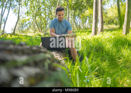 Mann mit Laptop sitzen auf Baumstamm in der Natur mit Tablet Stockfoto