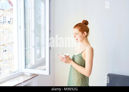 Portrait von nachdenklich Frau mit Schale Milchkaffee in der Nähe von geöffneten Fenster Stockfoto