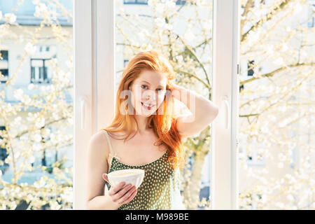 Porträt der rothaarige Frau mit Schale, weiß Kaffee vor Fenster Stockfoto