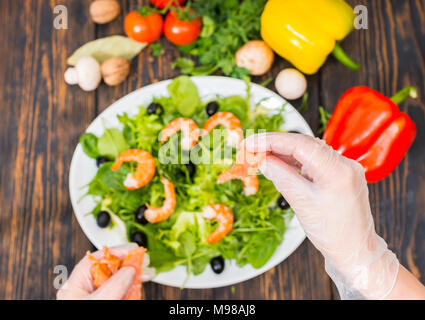 Hohe Betrachtungswinkel von Gemüse, Hände in Handschuhe holding Lachs oben Mix aus Salatblättern, Garnelen und Oliven in weiße Platte auf hölzernen Schreibtisch Stockfoto