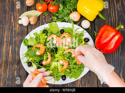 Hohe Betrachtungswinkel von Gemüse, Hände in Handschuhe legt Lachs auf Mix aus Salatblättern, Garnelen und Oliven in weiße Platte auf hölzernen Schreibtisch Stockfoto