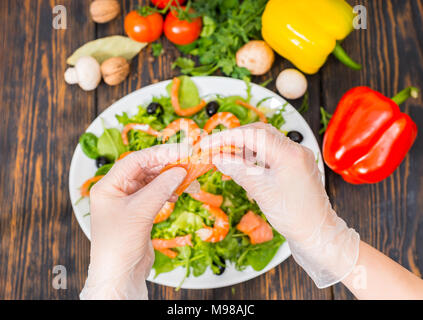 Hohe Betrachtungswinkel von Gemüse, unkenntlich Hände in Handschuhe holding Lachs oben Mix aus Salatblättern, Garnelen und Oliven in weiße Platte auf Holz Stockfoto