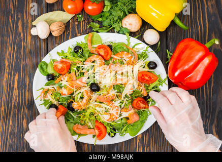 Hohe Betrachtungswinkel der unkenntlich Hände in Handschuhe legt aus Tomaten im Salat mit Mix aus Salatblättern, Garnelen, Oliven und Dijon Senf, Dressing Stockfoto