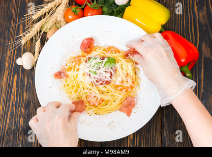 Hände in Handschuhe anziehen Jamon in Spaghetti Pasta mit Garnelen, geriebenem Käse, Tomatensoße und Basilikum in weißer Teller mit Gemüse auf Holz des Umgeben Stockfoto