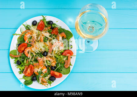 Hohe Betrachtungswinkel von leckeren Salat mit Garnelen, geriebener Käse, Mix aus Salatblättern, Oliven in weiße Platte in der Nähe von Glas mit Wein auf Blau Holz- Tabl Stockfoto