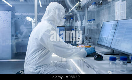 In einem sicheren Hohe Labor Wissenschaftler in einem Overall, die Durchführung einer Forschung. Biologe analysiert die Ergebnisse auf einem Computer. Stockfoto