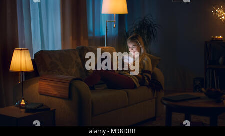 Am Abend schöne junge Frau liegt auf der Couch und Smartphone verwendet. Zimmer aussieht, warm und gemütlich. Stockfoto