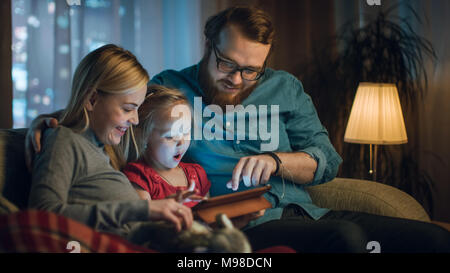 Vater, Mutter und Tochter lesen Kinderbuch auf einem Sofa im Wohnzimmer. Es ist Abend. Stockfoto