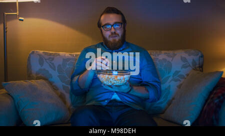 Porträt der Mann sitzt auf einem Sofa in seinem Wohnzimmer, Popcorn essen und Fernsehen. Standleuchten eingeschaltet sind. Stockfoto