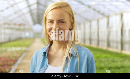 Der schöne Blonde Gärtner steht lächelnd in einem Gewächshaus voller bunter Blumen. Stockfoto