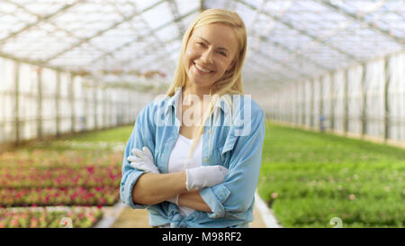 An einem sonnigen Tag schöne Blonde Gärtner steht lächelnd in einem Gewächshaus voller bunter Blumen. Stockfoto