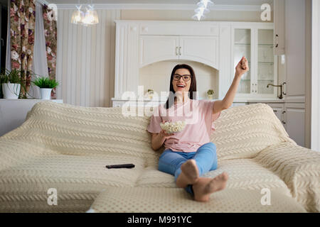 Eine brünette Mädchen mit Brille mit Popcorn vor dem Fernseher sitzen auf dem Sofa im Zimmer. Stockfoto