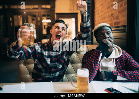 Hübscher junger Mann mit seinem Bier und gerne schreien auf dem TV-Bildschirm, Fußball Spiel mit seinem umgekippt afro-amerikanische Freunde am Schnittlängengetriebe Stockfoto