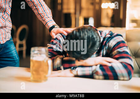 Menschen, Freizeit, Freundschaft und Party Konzept - Mann mit Bier seinen betrunkenen Freund schlafen auf dem Tisch an der Bar oder pub Aufwachen Stockfoto