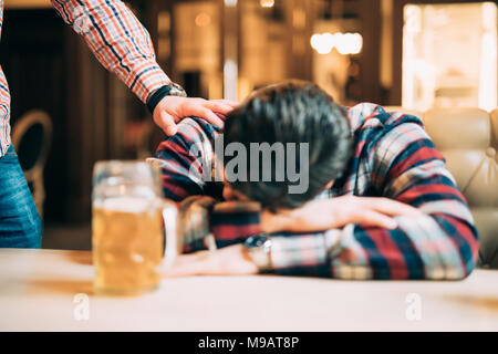 Junger Mann in Freizeitkleidung ist schlafen in der Nähe der Becher Bier auf einer Tabelle in ein Pub, ein anderer Mann ist wecken ihn auf. Stockfoto